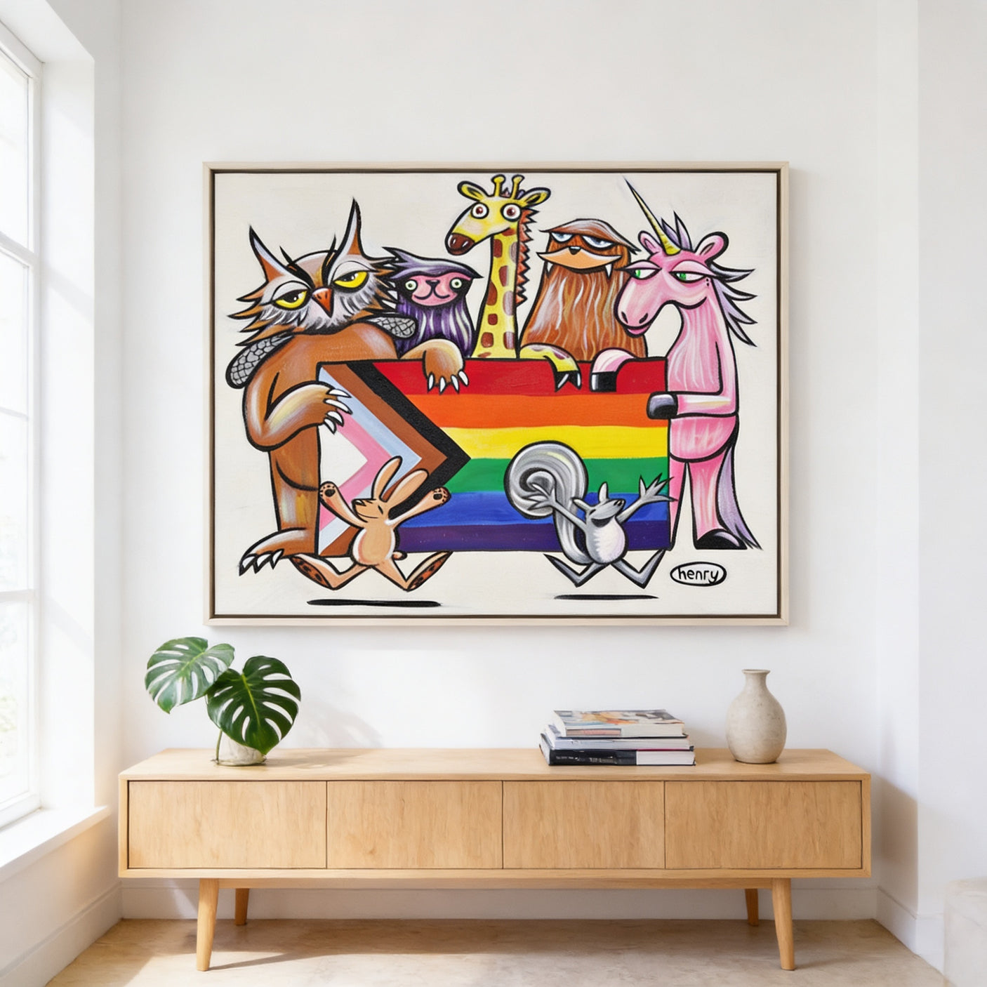 a vibrant and colorful painting of a group of animals, including a cat, giraffe, unicorn, and rabbit, all holding a rainbow flag. The painting is displayed on a white wall above a wooden cabinet, which has a plant and some books on it.